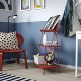 Red KRÖNIKÖR shelf with books, speaker, & chair with pillow against blue wall, coat rack nearby.