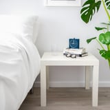 White LACK side table beside bed with glass, books, clock, plant.
