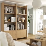Wooden storage unit with open and closed shelves, white decor items, lamp, and windows. Modern minimalist living room.