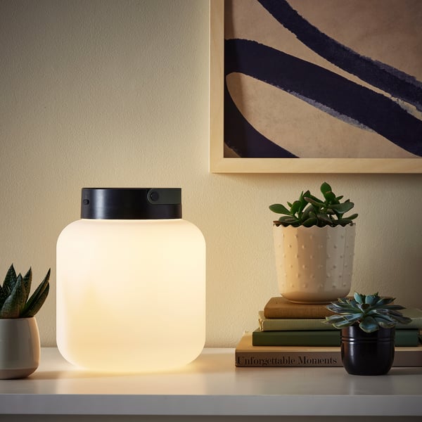 A modern living space with a LÄNSPORT portable lamp glowing warmly, surrounded by plants and books.