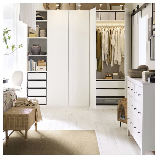Modern white bedroom with beige rug, wicker bench, and open closet showing clothes, shelves, and drawers.