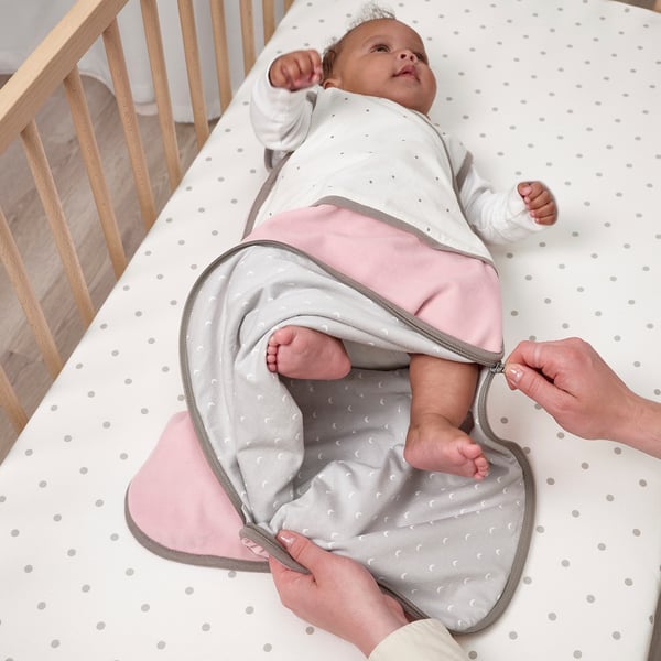 Adult zips baby into a cozy sleep bag on a white polka-dotted mattress.