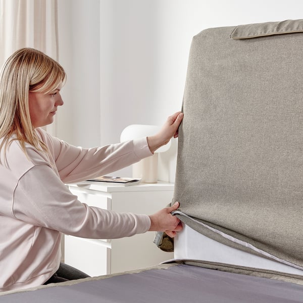A woman adjusts a gray fabric-covered divan bed with a sloped headboard and extra cushions in a well-lit room.