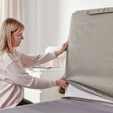 A woman adjusts a gray fabric-covered divan bed with a sloped headboard and extra cushions in a well-lit room.