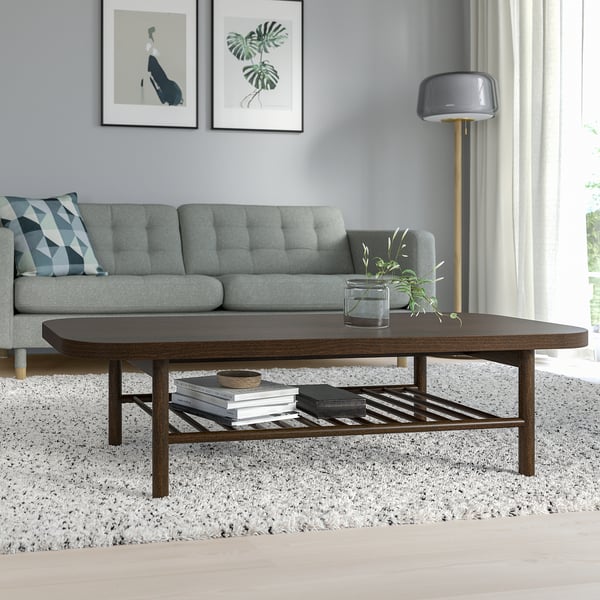 Modern living room with LISTERBY dark brown wooden coffee table, gray sofa, and tall floor lamp. Table has a lower shelf with books and a plant.