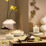 White vanilla candles in glass, varied sizes, wooden table with orchids.