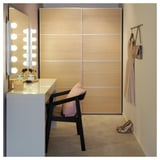 Modern vanity setup with lighted mirror, white table, black chair, and wooden closet. Small accessories laid out.