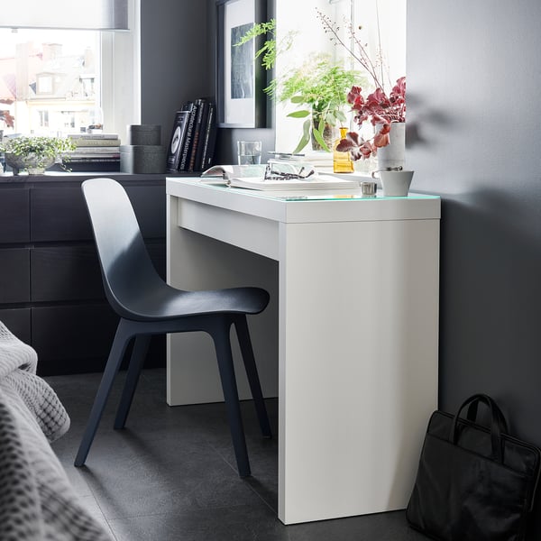Modern black chair beside a white desk with glass top, featuring a neatly arranged workspace with decor and books in a stylish room setting.