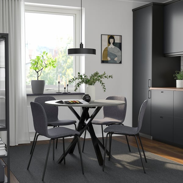 Modern dining set with round table, grey chairs, flowers, and candles. Large window and dark cabinets in background.