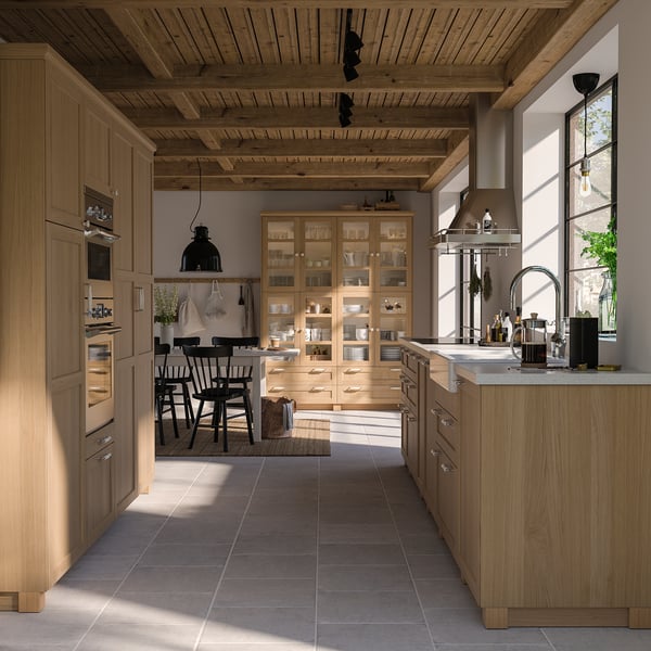 Modern kitchen with light oak cabinets, black chairs, and large window. Wooden ceiling, stainless appliances, and glass display unit.