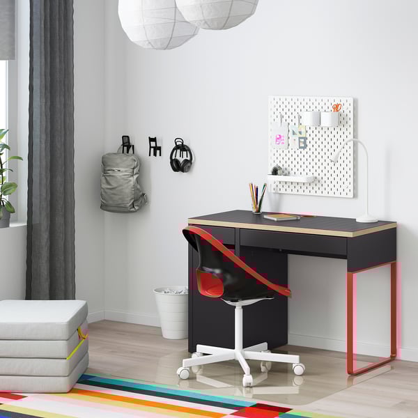 Modern workspace with black MICKE desk, red chair, wall storage, grey backpack, white ottoman, colorful rug.