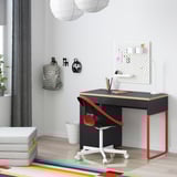 Modern workspace with black MICKE desk, red chair, wall storage, grey backpack, white ottoman, colorful rug.