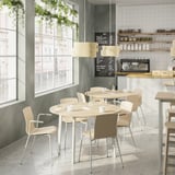 Modern café with round tables and chairs. Light wood tables, 4-6 seats, natural light and plants.
