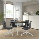 MITTZON conference table with black top, white legs, and black chairs for small meetings. Cable tray, window, plants, and storage cabinet.
