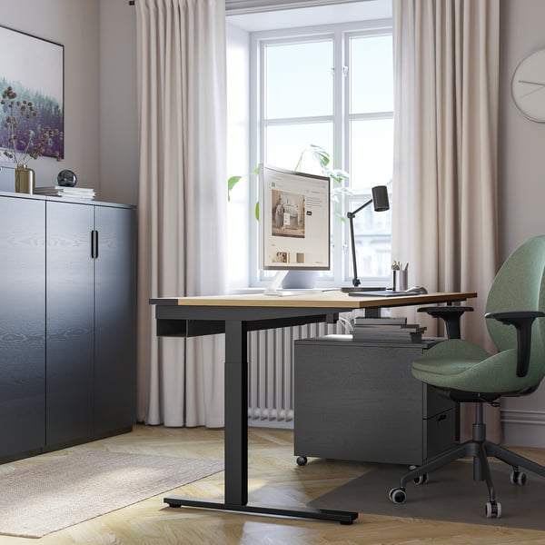 Modern home office with MITTZON height-adjustable desk, green chair, computer, and storage cabinet. Against window with curtains.