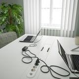 White desk with two laptops, power strips, and cables. Plant and chair nearby.