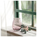 Purple recycled glass vase on wooden window sill, holding pink flowers, next to green leaves.