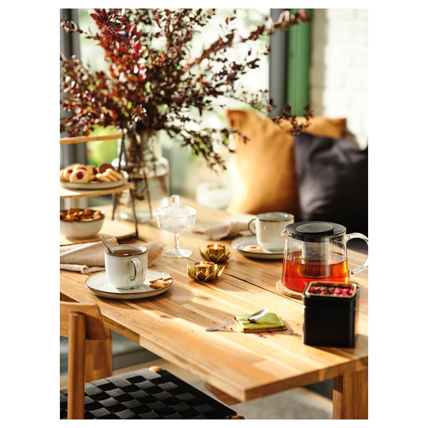 A cozy wooden table set with tea cups and treats, featuring a glass teapot and gold accents, surrounded by chairs.