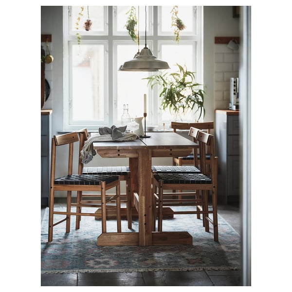 Rustic wooden dining set with acacia table and 4 woven chairs near large window; cozy decor with plants and natural textiles.