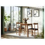 Modern dining set with round wooden table and chairs near bright window. White brick wall with art.