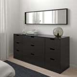 Modern black NORDLI chest with mirror, lamp, flowers, and books. Beside bed with white blanket and gray rug.