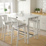 Bright kitchen nook with white NORDVIKEN bar table and stools. Table has drawer, tea set up top. Large window, plants, rug.