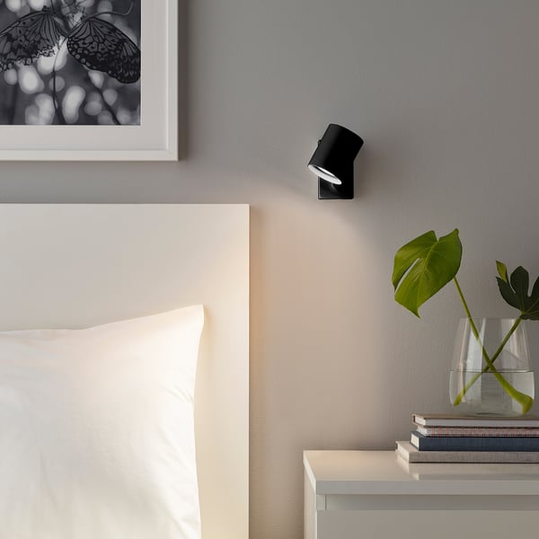 NYMÅNE wall lamp in black close to a bed, with a vase, and art. Small, sleek design.