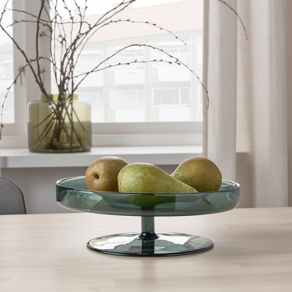 A glass bowl holds three pears on a wooden table. Behind it, a green vase with branches sits near a window.