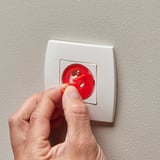 A hand pushes a red childproof safety plug into a white wall socket, ensuring electrical safety for children.