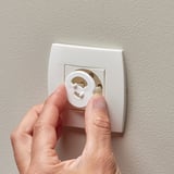 Hand inserts white safety plug into wall socket to prevent access.