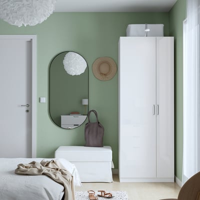 Minimalist bedroom with white PAX wardrobe, mirror, and green walls. Bed with white linen and beige blanket. Decor includes lamp, hat, and bag.
