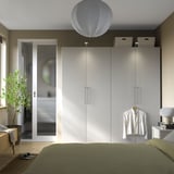 Modern bedroom with a tall white PAX wardrobe and mirror. Sleek sliding doors and a hanging shirt highlight its size and minimalist design.