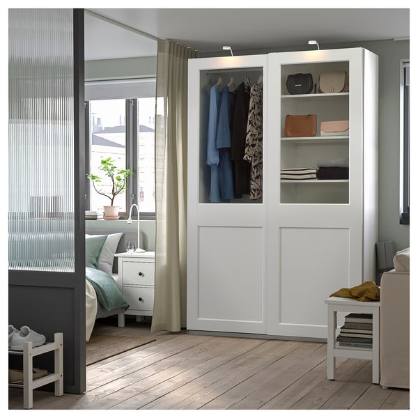 A white modern bedroom with a PAX wardrobe featuring glass doors, displaying clothes and accessories. The room includes a bed and a window.