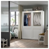 Modern bedroom with white PAX wardrobe showing clothes, a bed, nightstand, and window with curtains.