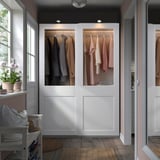 A white PAX wardrobe with glass doors displaying colorful clothes. It stands between windows and another wardrobe, offering ample storage.