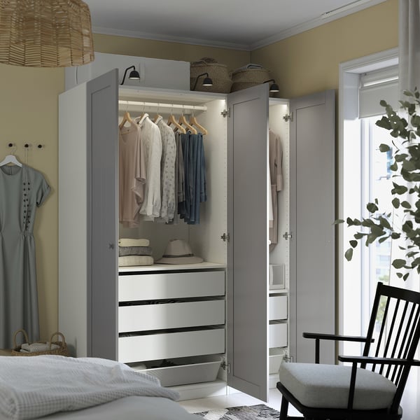 Open wardrobe with clothes, hats, and baskets on top; bed with blanket and chair nearby.