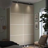 A modern bedroom a tall beige PAX wardrobe with sliding doors, soft lighting, a side table with a cushion, and a potted plant.
