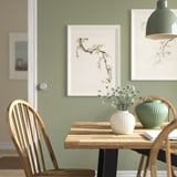 Cozy dining area with wooden table, PELARBJÖRK vase with flowers, under modern lamp, sage green wall with pictures.