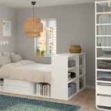 Minimalist bedroom with PLATSA bed, storage, and books. Light wood floors, grey walls, floral rug. Large window, warm pendant lamp.