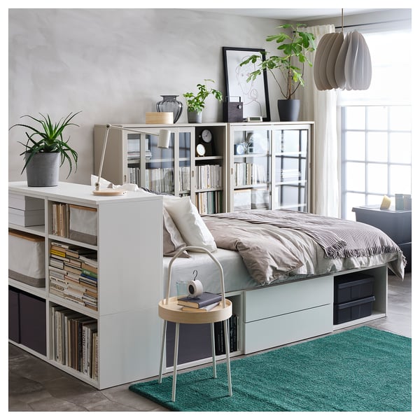 Modern bedroom with a white PLATSA bed, grey linens, and built-in storage. Decorated with plants, books, and a small round side table.