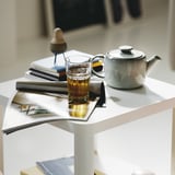 A glass of tea atop a magazine on a white table, with a ceramic teapot beside it.
