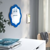 Blue-framed, wavy mirror on wall, reflecting interior. Beside white drawer with books and decorative tray.
