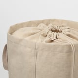 Cylindrical beige fabric laundry tote with drawstring closure. Handles on either side for carrying. Seamless design, ready for laundry duties.
