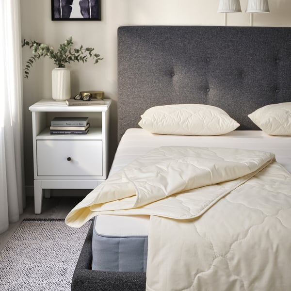 Bedroom: twin bed with RÅDHUSVIN duvet in natural color. Grey headboard, white pillows, nightstand with books and vase.