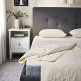 Bedroom: twin bed with RÅDHUSVIN duvet in natural color. Grey headboard, white pillows, nightstand with books and vase.