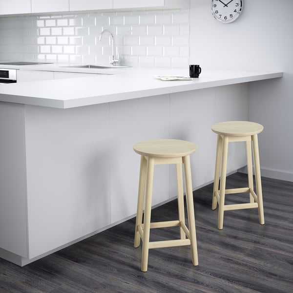 Two light wooden bar stools sit before a white kitchen counter with a sink and clock.