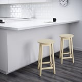 Two light wooden bar stools sit before a white kitchen counter with a sink and clock.