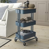 Blue RÅSKOG trolley near couch with coffee maker, mugs, towels, headphones, books.