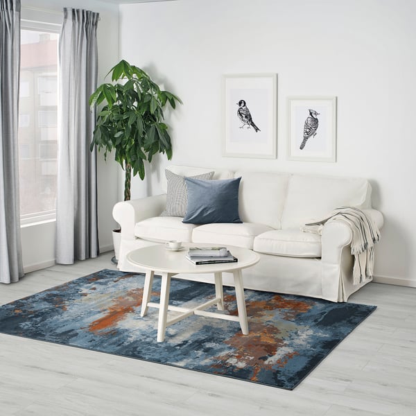 Modern living room with white sofa, abstract rug, and tall plant. Artwork and coffee table on rug.