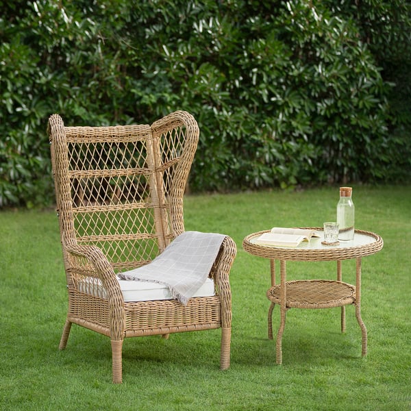 Wicker chair & side table on lawn, rattan, cushion, blanket, glassware, book, green bushes.